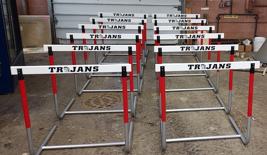 Twelve custom track hurdles with red uprights and white TROJANS crossbars