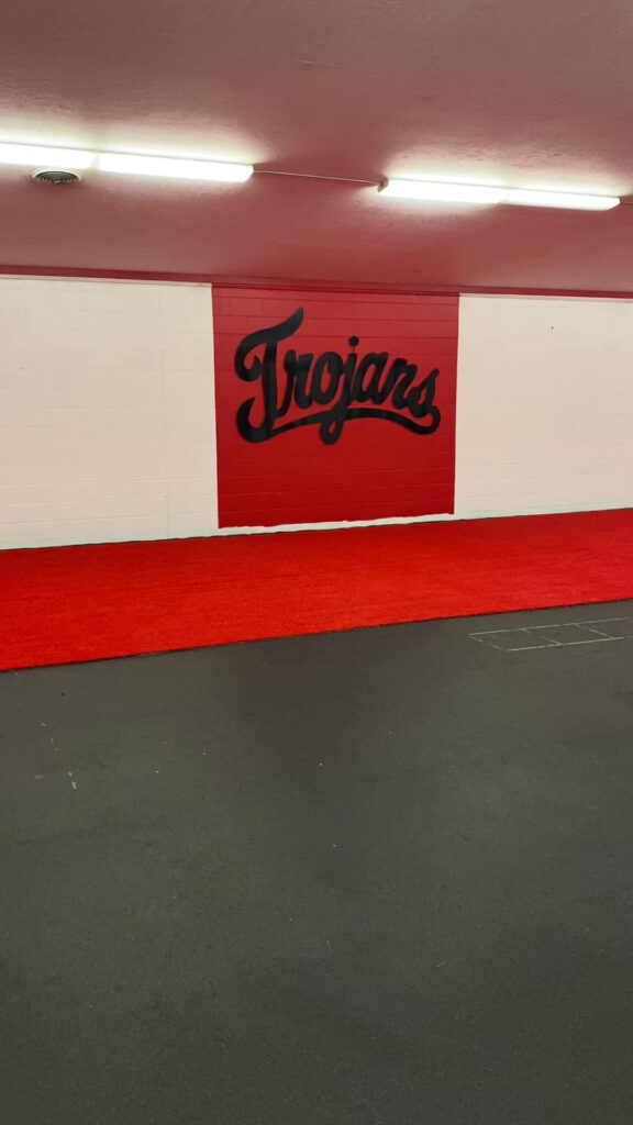 Remodeled training facility hallway with a black Trojans script logo on a red wall and new red turf